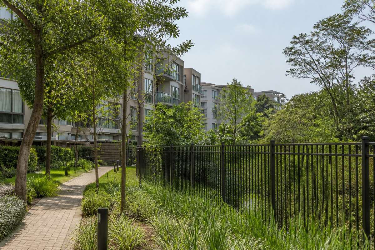 Área verde protegida em um condomínio residencial moderno, com árvores, caminhos e edifícios ao fundo.